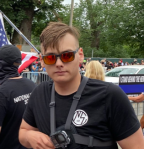 Photo of Chris Hood at a rally wearing mirrored orange sunglasses and wearing a black NSC t-shirt