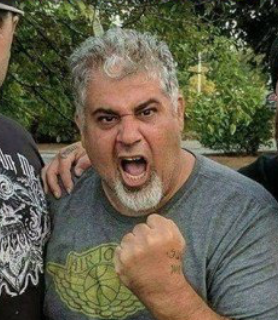 Photo of Kenny Lizardo, he has a goatee and greying hair, his mouth is open and he is holding a fist up in front of his chest. 