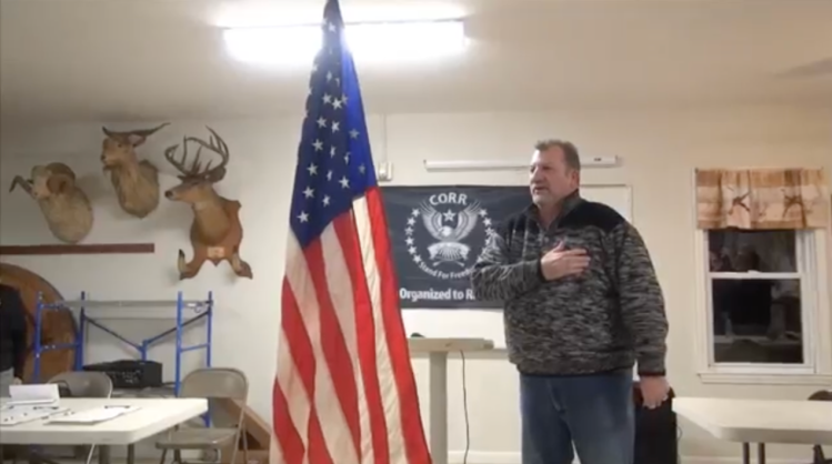 John McCaul stands in front of the American flag with his right hand over his heart. He is wearing a gray jacket and jeans. Behind him is the CORR flag hanging on the wall and the heads of three taxidermied animals.