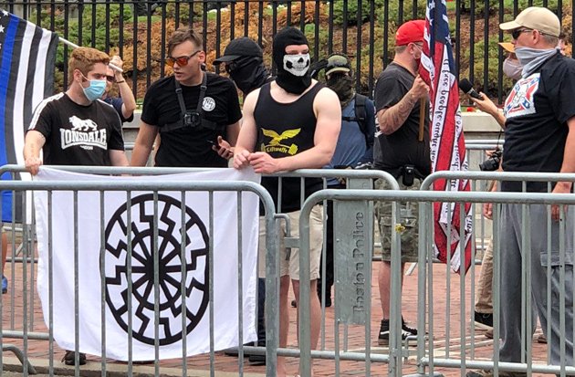 3 members of NSC including Chris Hood hold a Sonnenrad flag (black and white); in the background, US and thin blue line flags and masked white supremacists
