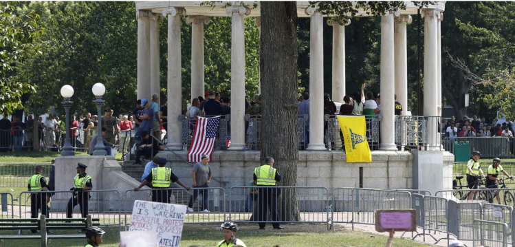 Boston Common bandstand hung with Gadsden and US flags, protected by Boston Police and metal barriers; counterprotesters visible in the background