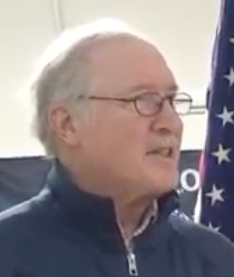 Headshot of pastor Mike Johnson. He is a white man in his mid-60s, with gray hair and glasses. He is standing in front of an American flag with the CORR banner in the background. He is wearing a blue zip up jacket.
